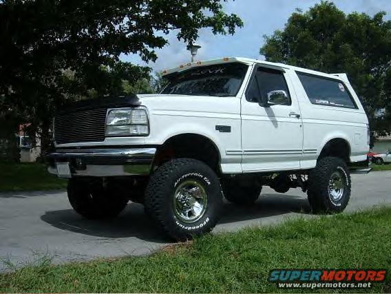White lifted SUV parked on a street, trees overhead. Has aftermarket grille and tires