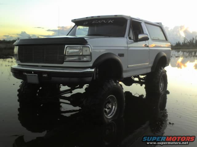 White lifted Ford Bronco with large tires sitting in shallow water, sun setting in the background