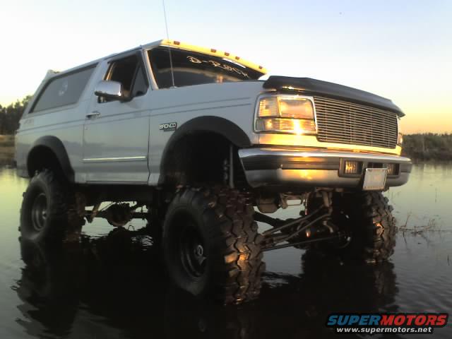Lifted 1995 Ford Bronco with large tires driving through shallow water at sunset