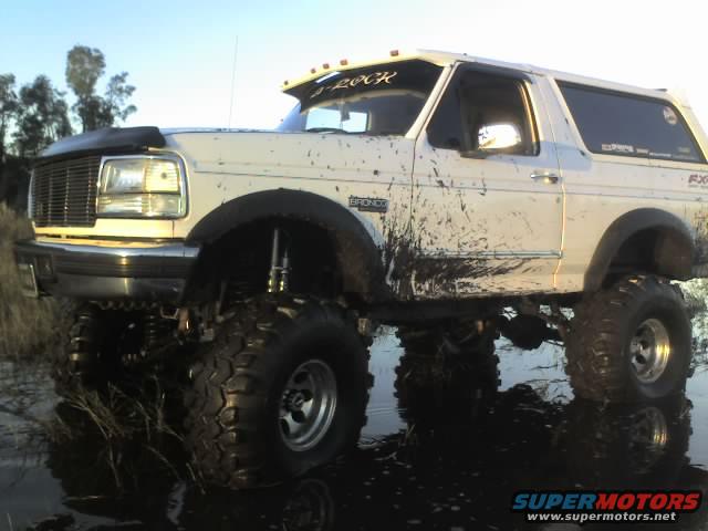 White 1995 Ford Bronco covered in mud, lifted with large tires, in a muddy puddle
