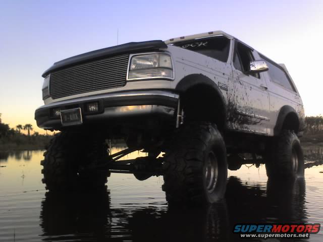 Lifted white Ford Bronco with large tires driving through shallow water. Mud splattered on the side