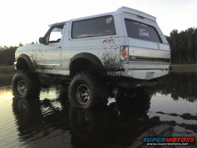 White 1995 Ford Bronco with lifted suspension and large tires driving through mud