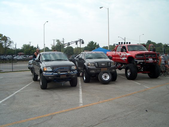 Three lifted pickup trucks parked in a lot, one red with flames, one gray with spare tires