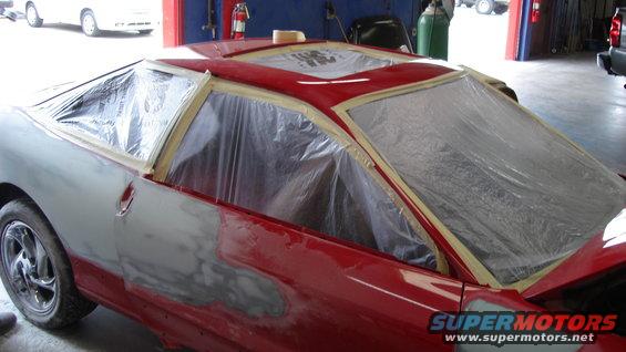 Car bodywork with windows taped off for painting. Red paint is visible on the roof and rear, with gray primer on the front fender and door