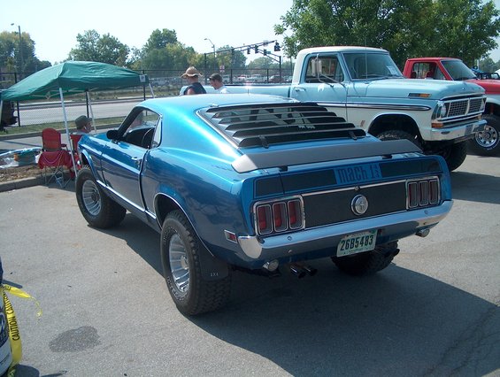 Blue 1969 Ford Mustang Mach 1 lifted with off-road tires, parked next to a light blue and white pickup truck