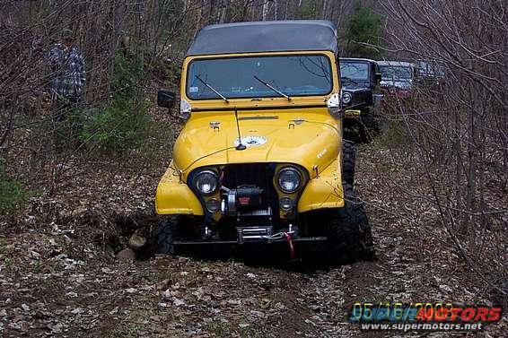Yellow Jeep CJ-5 with winch on front bumper, driving on a dirt trail with other Jeeps behind it