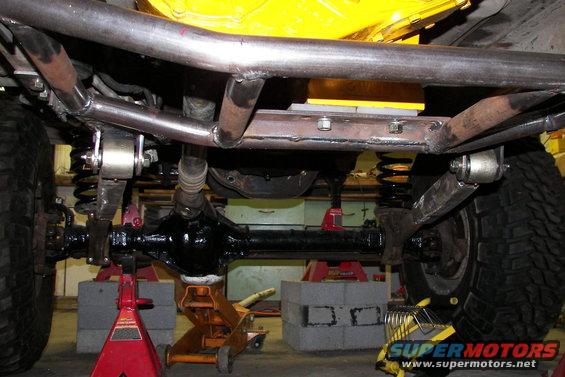 Underbody view of a vehicle showing suspension components, differential, and frame modifications. Vehicle is supported by jack stands