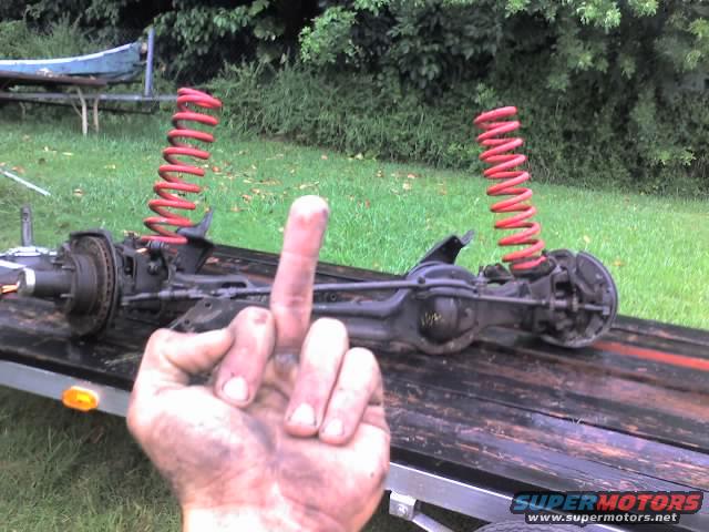 Vehicle axle assembly with red coil springs on a trailer, hand giving the middle finger in foreground