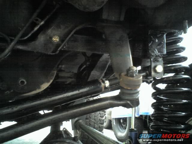 Underside view of a vehicle's suspension with a large coil spring and metal components. Text reads "SUPERMOTORS.NET"