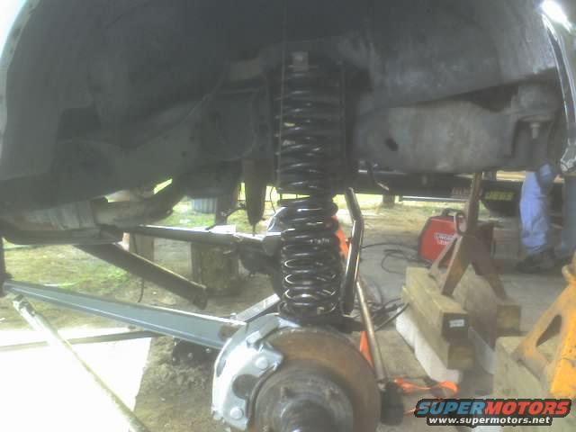 Close-up of vehicle suspension components, including a coil spring and shock absorber, with a welder in the background