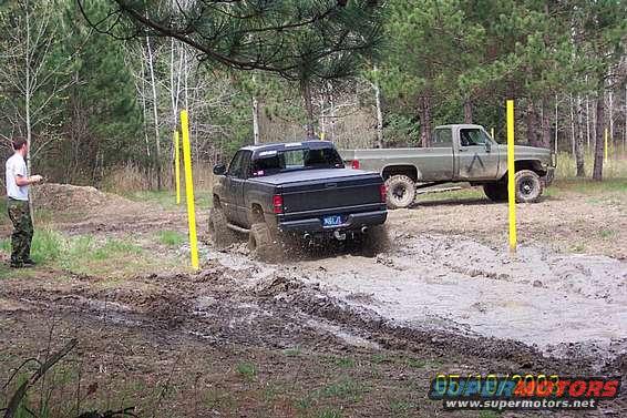 Black pickup truck driving through mud, green pickup truck parked behind it. Person standing to the left