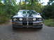 1966 Ford Mustang - A gray Ford Mustang with racing stripes is parked on a paved surface surrounded by trees.