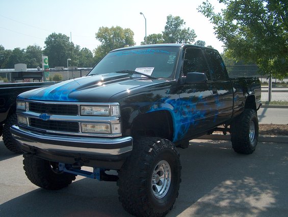 Black pickup truck with blue flame graphics on hood and side, lifted with large tires
