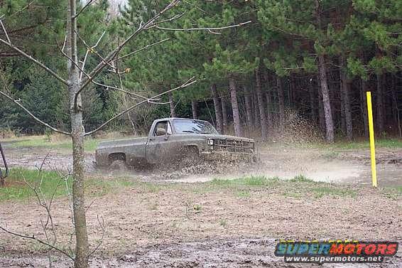 Olive green pickup truck driving through mud, splashing water. Trees and a yellow pole in background