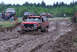 1989 Ford Bronco - A red SUV navigates a muddy track during an off-road event, with other vehicles and spectators in the background.