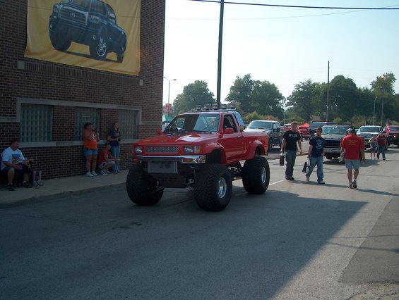 Red lifted pickup truck with large tires, custom grille, and roof lights, seen from a 3/4 front angle