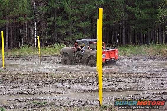 Off-road vehicle with orange bed stuck in deep mud, surrounded by trees