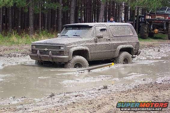 Mud-covered Chevrolet K5 Blazer stuck in mud, front 3/4 view. Visible grille and headlights suggest 1980s model