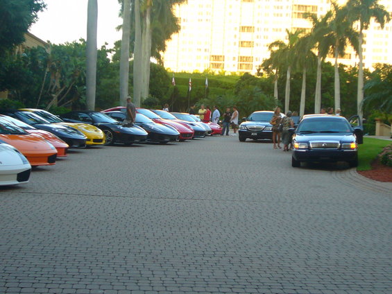 Several sports cars and sedans parked in a lot with palm trees and a building in the background