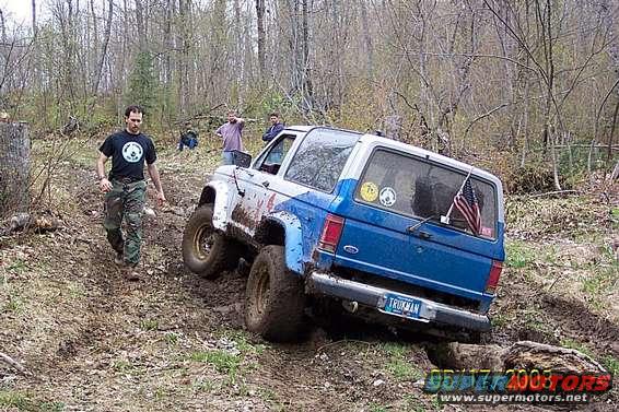 Blue and white SUV with "TRUMAN" license plate, tilted on muddy terrain