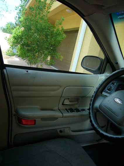 2001 Ford Crown Victoria interior showing door panel, steering wheel with Ford logo, and side mirror. Exterior view through window shows a garage and tree