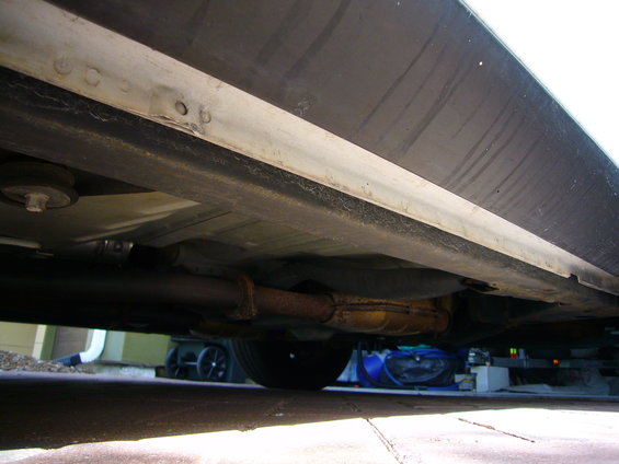 Underside view of a car, showing the exhaust pipe and part of the undercarriage. Some items are visible in the background