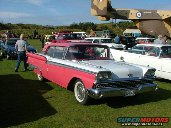 1959 Ford Fairlane 500 with pink and white two-tone paint, parked on grass with other cars and a plane in the background. License plate reads GSK 486