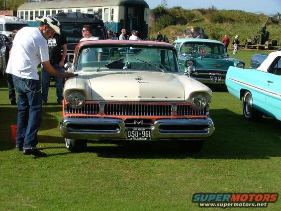 Front view of a cream and orange 1957 Mercury Montclair with a man cleaning it. Other vintage cars and a train car are in the background