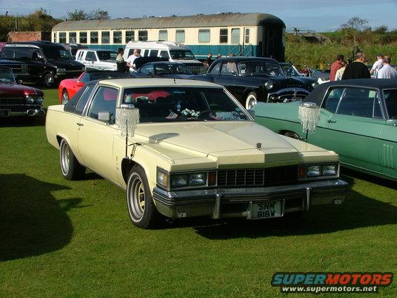 Cream 1977 Cadillac Coupe DeVille with a black vinyl roof, parked on grass. Front view shows chrome grille and bumper