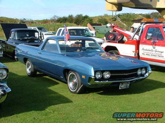Blue 1970 Ford Ranchero parked on grass with a tow truck and black pickup in background