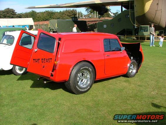 Red panel van with "THE BEAST" painted on the rear door, parked on grass next to a white vehicle. Custom wheels and open hood