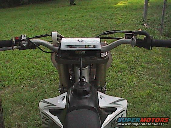 Motorcycle handlebars with a silver crossbar pad and black grips. White and black fuel tank visible below