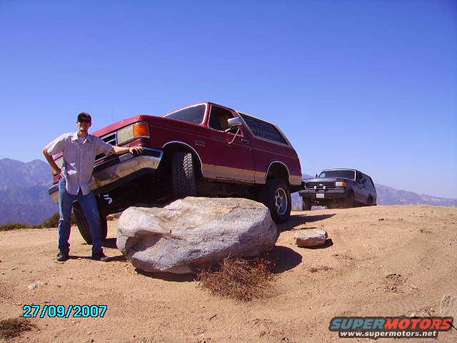 Red Ford Bronco on a rock, driver standing next to it. Dark SUV in background