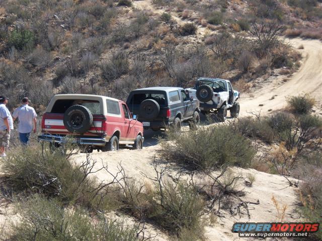 Three Ford Broncos driving on a dirt trail in a dry, hilly area. One red Bronco is in the foreground, followed by a dark Bronco, and then a white Bronco