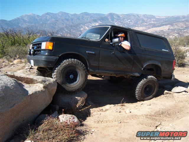 Black Ford Bronco climbing over rocks on a dirt trail with mountains in the background