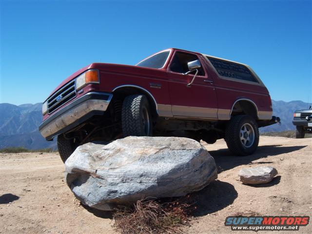 Red Ford Bronco with tan trim on a rock, another vehicle in background
