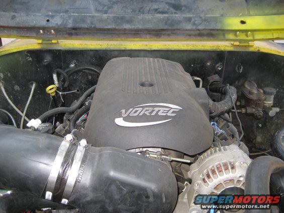 Vortec engine cover with air intake tube and alternator visible. Supermotors.net logo at bottom right