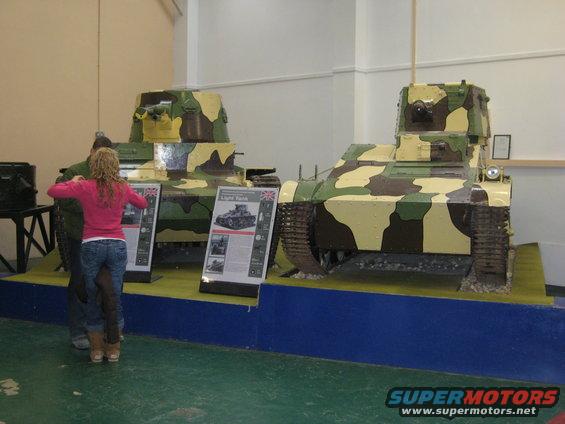 Two camouflaged tanks on display with informational signs in front of them