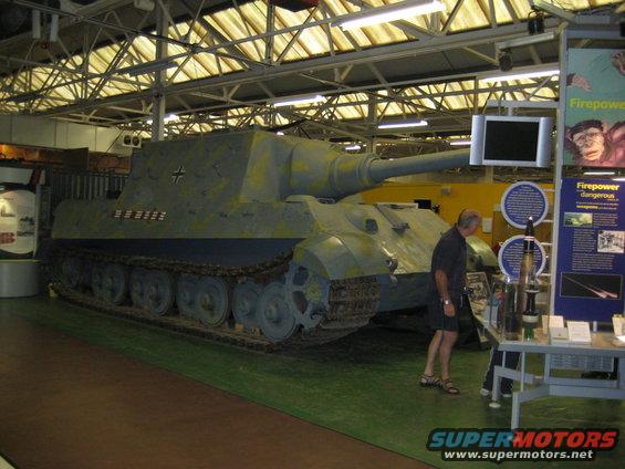 Jagdpanther tank destroyer with camouflage paint, viewed from the front-right. A man stands nearby looking at displays