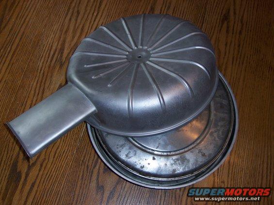 Metal air cleaner assembly with ribbed lid and rectangular intake pipe, resting on a wooden surface