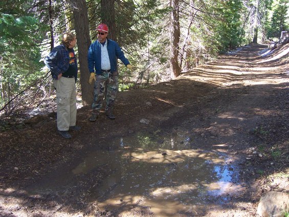 Two men stand on a muddy forest path with a large puddle in the foreground