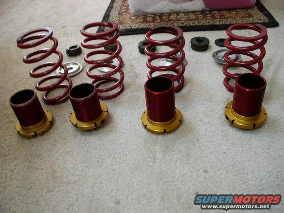 Four red coilover springs and red shock bodies with gold bases are laid out on a carpeted floor