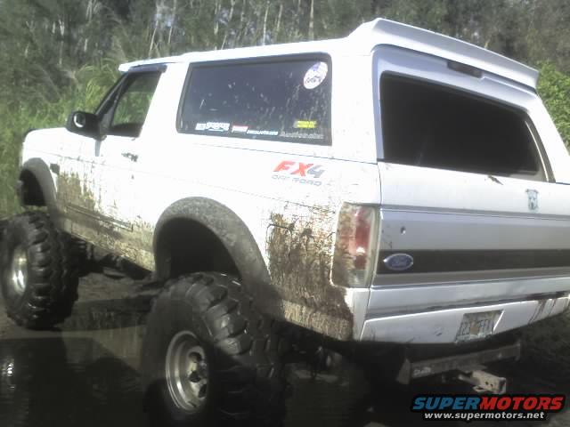 White 1995 Ford Bronco with large tires, covered in mud, parked in water