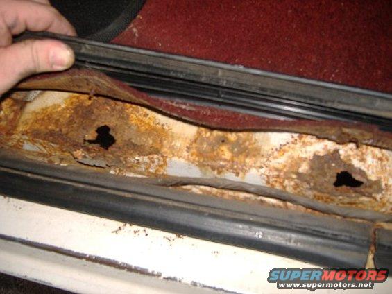 Door sill heavily rusted with holes, showing red carpet and black rubber seal