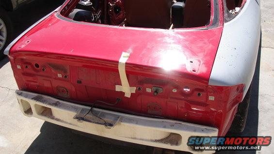 Car's red rear end with a white bumper. The trunk area is open, showing exposed metal and tape