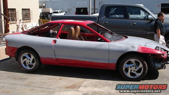 Primered and red car with roll cage, tan seats, and chrome wheels, side view
