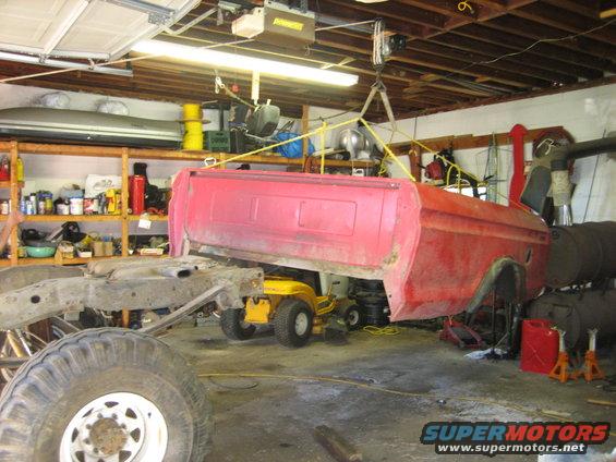 red truck bed lifted by hoist in garage, chassis and large tire in foreground