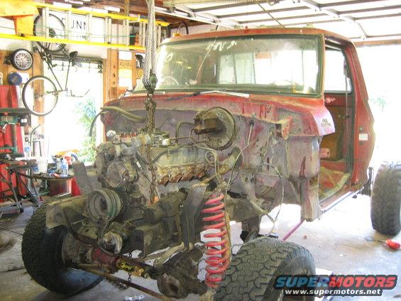 Red truck chassis with engine removed, lifted by a chain hoist. Visible are the front suspension, tires, and part of the cab