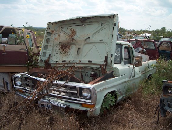 light blue pickup truck with hood open, parked in a junkyard next to a maroon SUV
