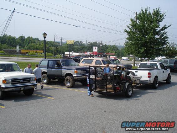 1987 Ford Bronco parked next to a white pickup truck towing an ATV on a trailer. Other vehicles and a gas station are in the background